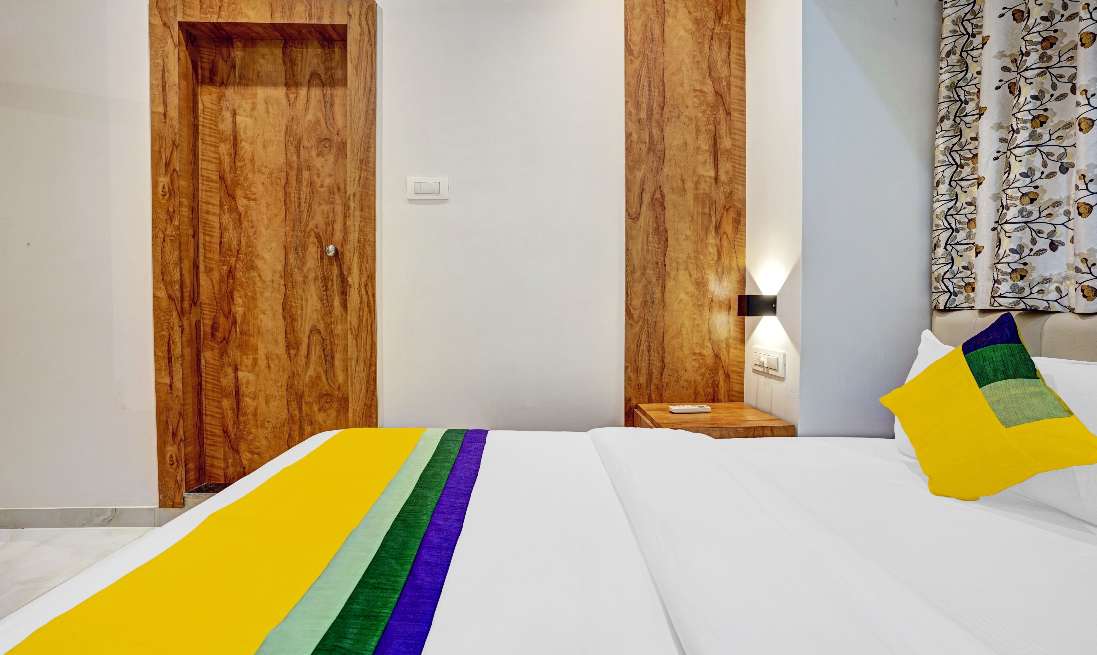 A hotel room with a wooden wardrobe, a bedside table with a lamp, and a bed with a colorful blanket.