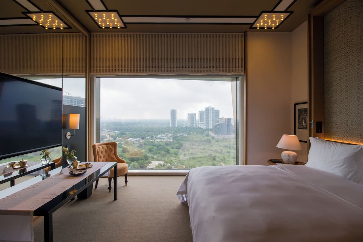 A hotel room with a large bed, a desk with a flat-screen TV, a chair, a nightstand with a lamp, and a large window offering a city view.