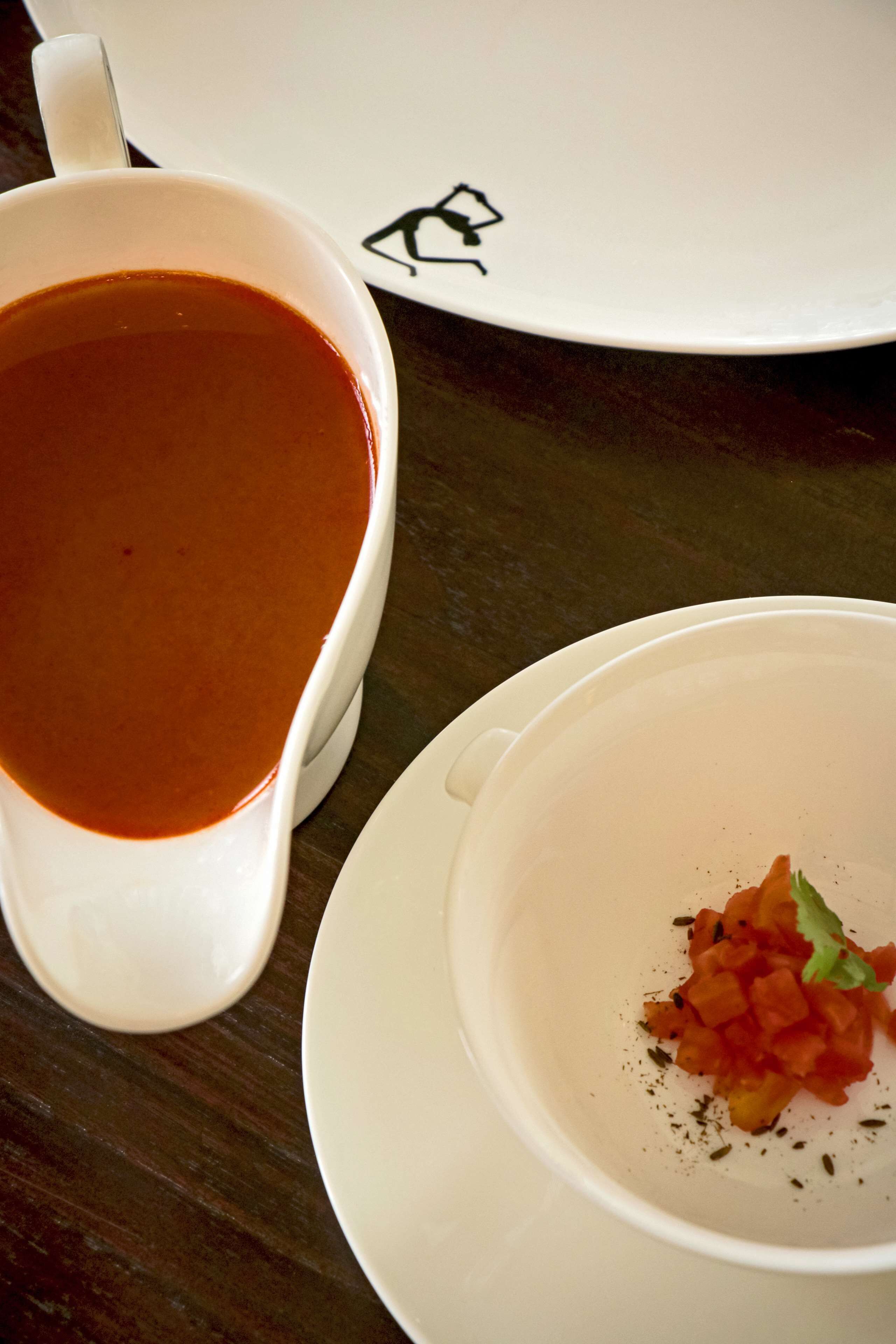 A white cup of tomato soup and a white bowl with diced tomatoes and herbs on a wooden table.