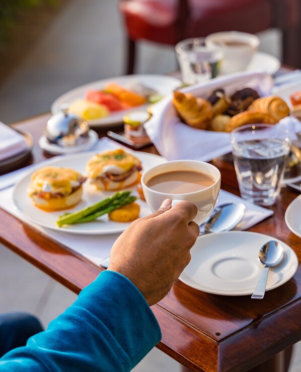 Una persona sosteniendo una taza de café en una mesa con alimentos para el desayuno, como huevos, espárragos y pasteles.