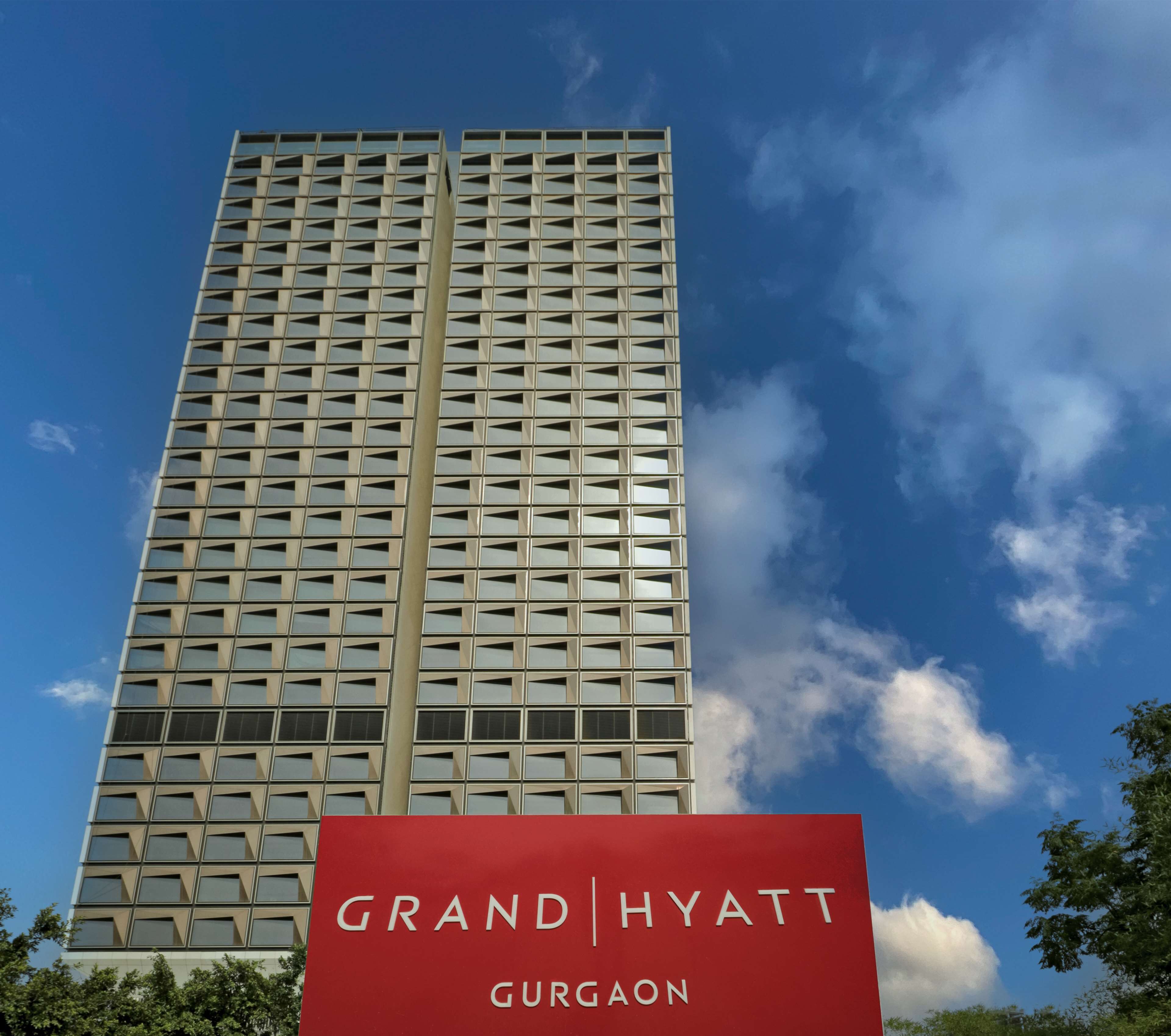 Un grand immeuble d’hôtel à la façade quadrillée, avec une grande enseigne rouge bien visible à sa base, sur laquelle on peut lire « Grand Hyatt Gurgaon ».