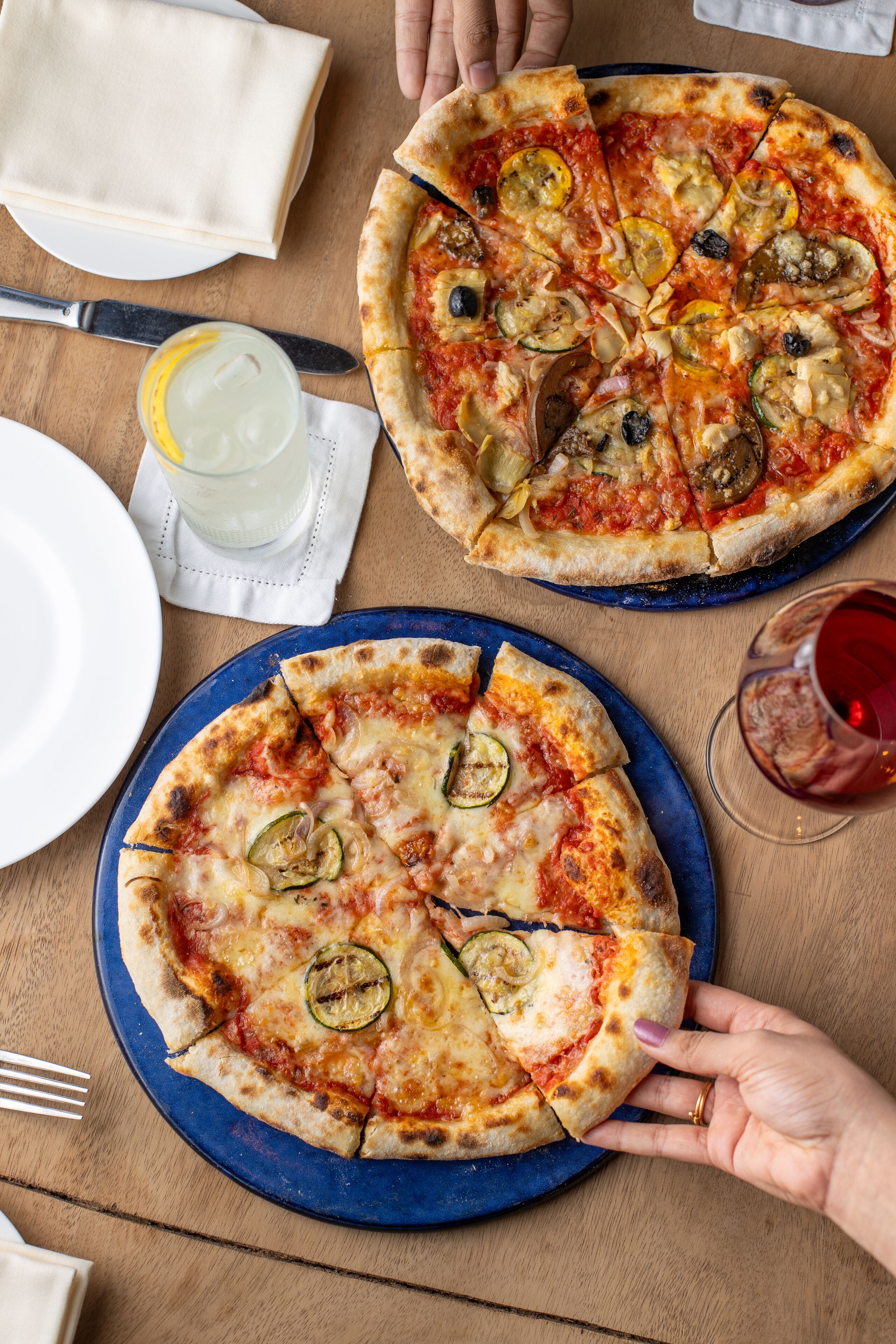 Dos pizzas sobre platos, un cuchillo, una servilleta y un vaso de agua en una mesa de madera.