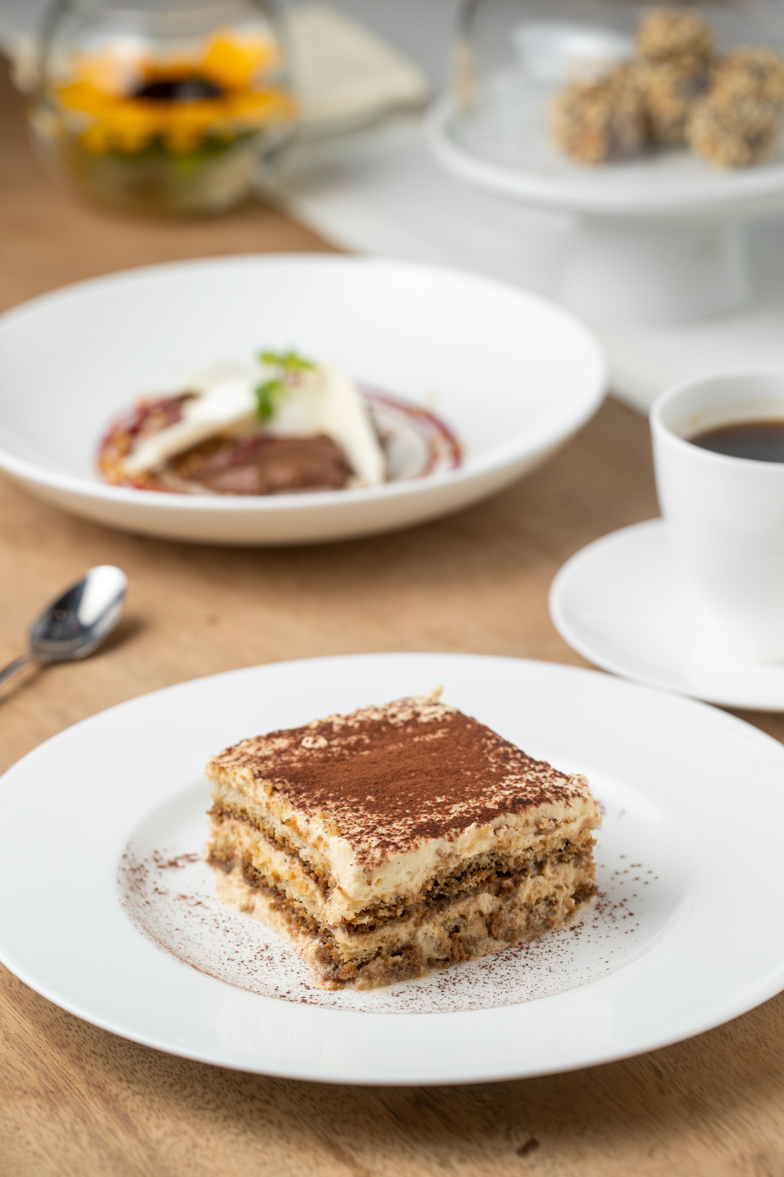 A slice of layered dessert on a plate, a cup of coffee, and a spoon on a wooden table.