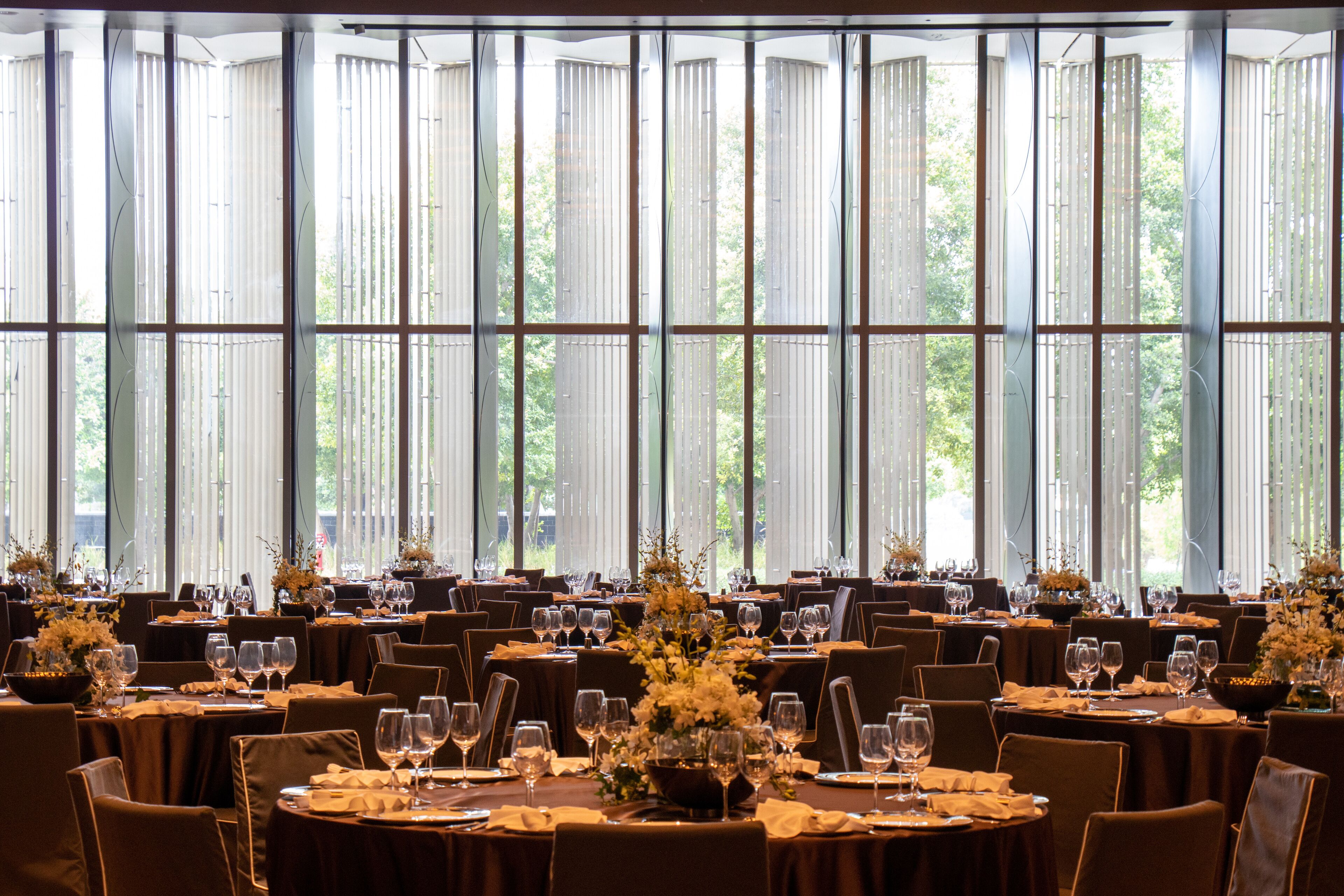 A large dining area with multiple tables set for a formal event, featuring tall windows and natural light.
