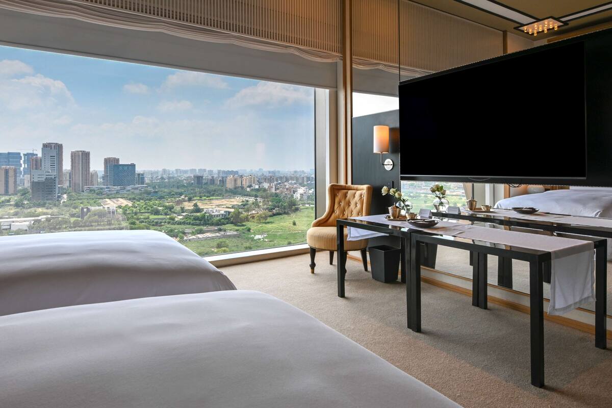 A hotel room with two beds, a desk, a chair, a TV, and a large window offering a city view.