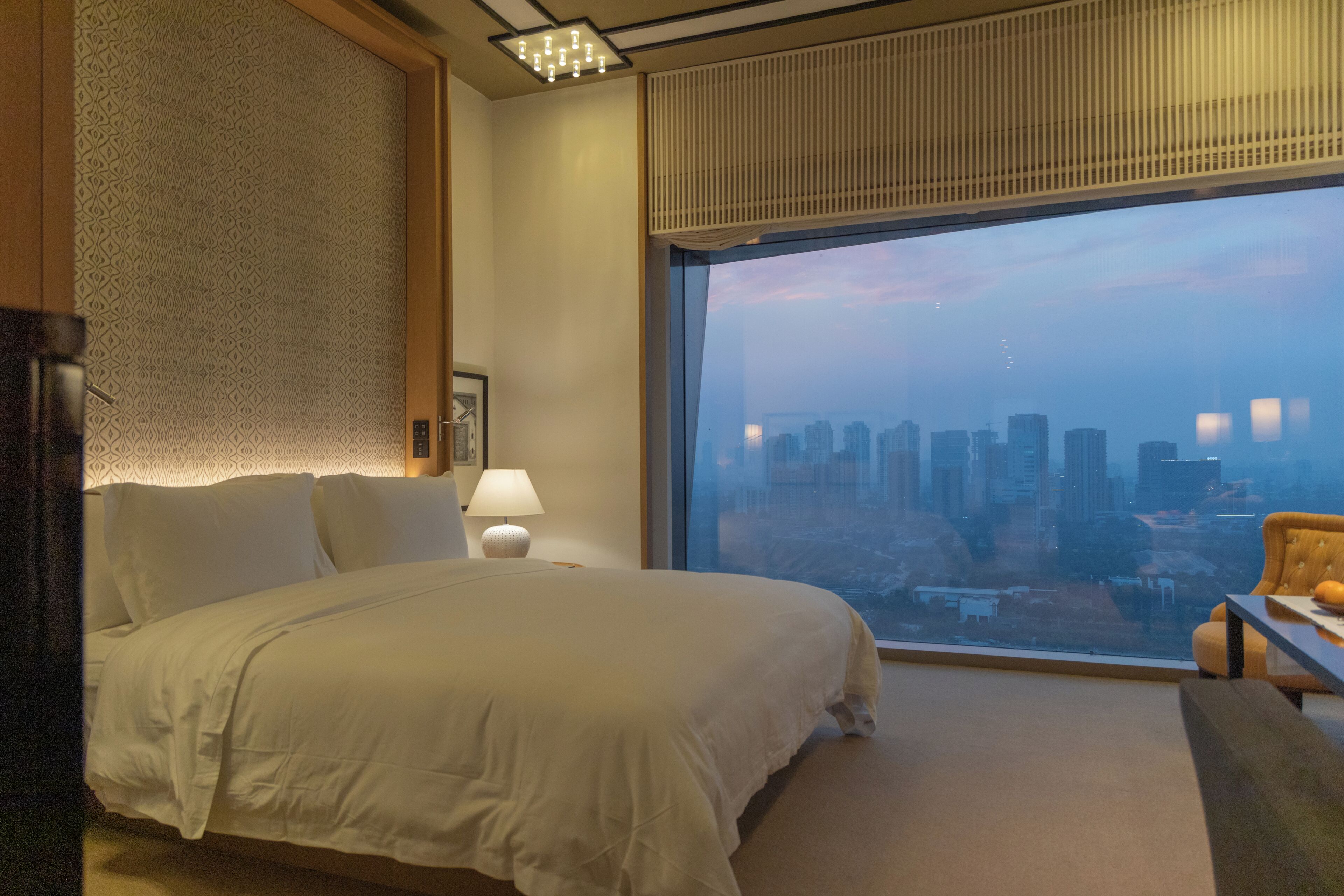 Hotelkamer met een groot bed, uitzicht over de stad, een raam met zonwering en een kleine tafel met een lamp.