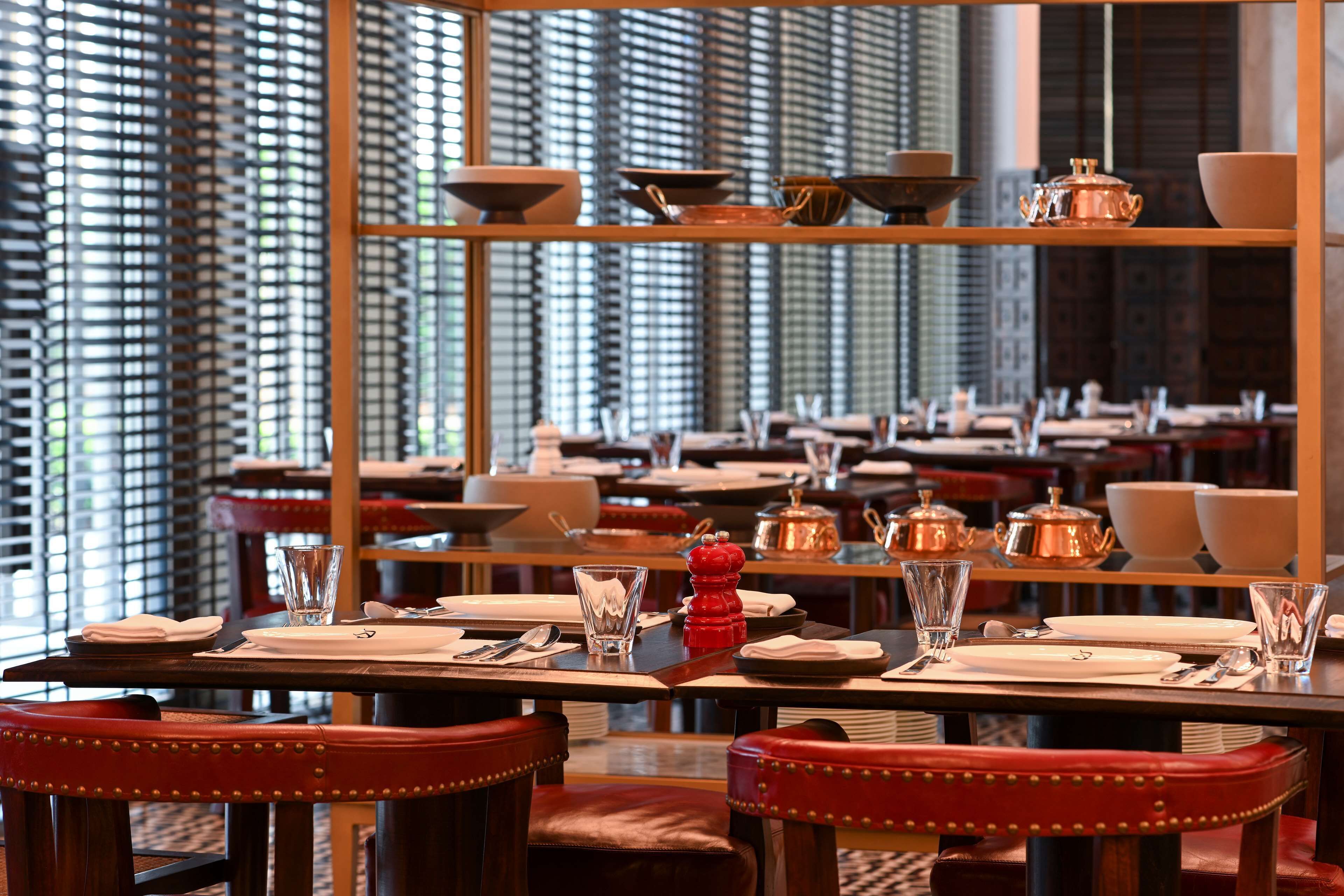 Un restaurant doté de nombreuses tables dressées pour le service, avec un intérieur moderne agrémenté de meubles en bois et d’une fenêtre décorative à motifs.