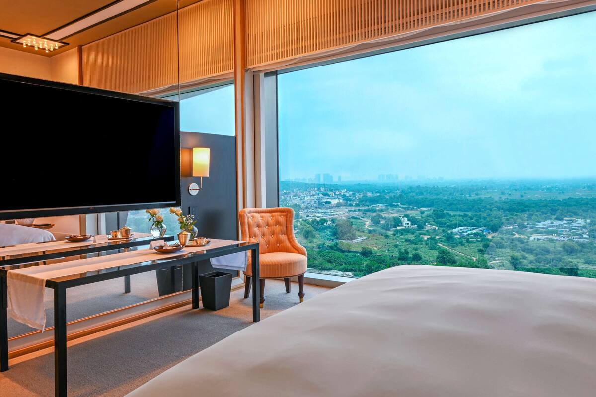 A hotel room with a large bed, a TV, a desk with a chair, and a view of a cityscape through large windows.