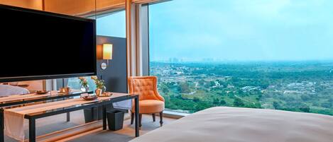 A hotel room with a large bed, a TV, a desk with a chair, and a view of a cityscape through large windows.