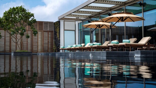 A modern pool area with lounge chairs and umbrellas, reflecting the building and tree. - Grand Hyatt Gurgaon (Gurugram)