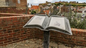 Un llibre obert amb text sobre un suport, amb un mur de maó de fons i una vista d'una ciutat i edificis a la distància.