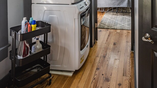 Pieni, käytännöllinen keittiö, jossa on jääkaappi, uuni ja siivoustarvikkeita sisältävä kärry. Avonaisen oven läpi näkyy sänky, jossa on vihreä tyyny.