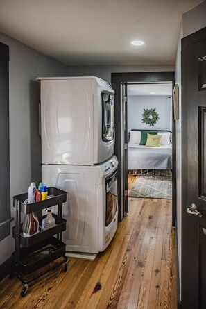 Una cocina compacta con refrigerador, horno y un carrito con productos de limpieza. Se ve una cama con un almohadón verde a través de una puerta abierta.
