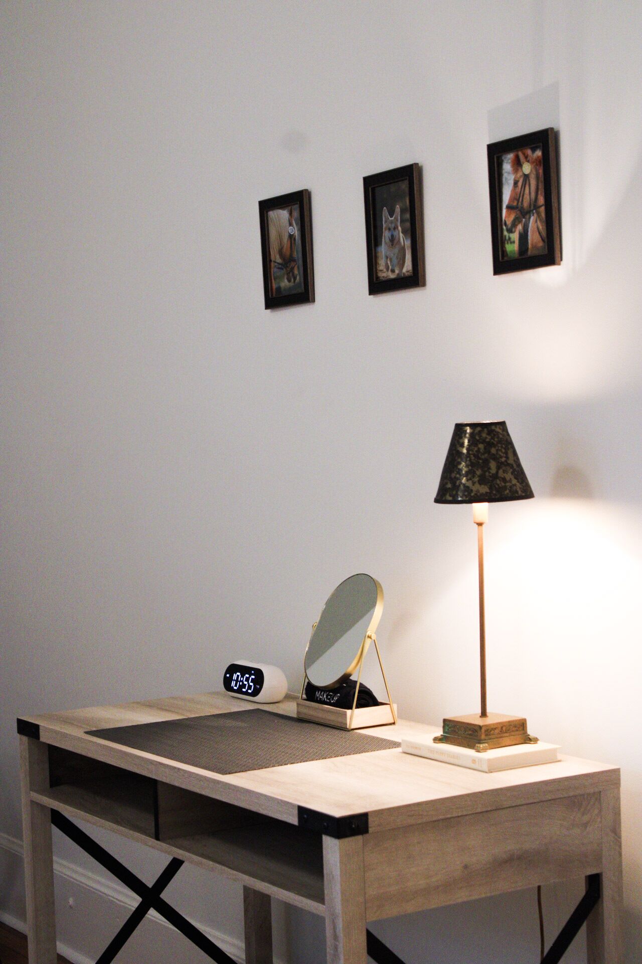 Un bureau avec une horloge numérique, une lampe et un miroir. Trois tableaux encadrés sont accrochés au mur, au-dessus du bureau.