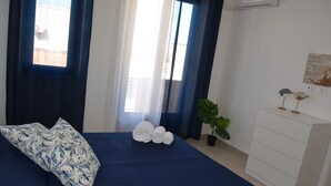 Una cama bien hecha con una colcha azul y una almohada decorativa, un juego de toallas dobladas, una planta en maceta y un tocador blanco con un adorno pequeño encima.