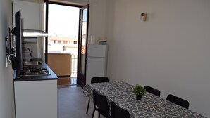 Una cocina compacta con estufa a gas, lavamanos y refrigerador. Hay una mesa de comedor con mantel estampado y cuatro sillas. Una pequeña planta en maceta está sobre la mesa.