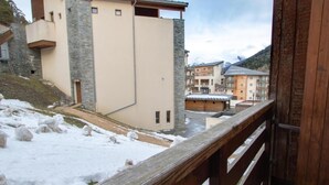 Un espacio exterior nevado con una terraza de madera, un edificio color beige con detalles de piedra y vistas a otros edificios al fondo.