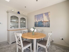 Un comedor con una mesa redonda de madera, dos sillas, un aparador con puertas de vidrio y un cuadro con barcos de vela en la pared.