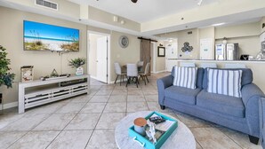 Un salón moderno con un sofá gris, una mesa de centro y un televisor que muestra una escena de playa. Hay una zona de comedor con sillas y una cocina al fondo.