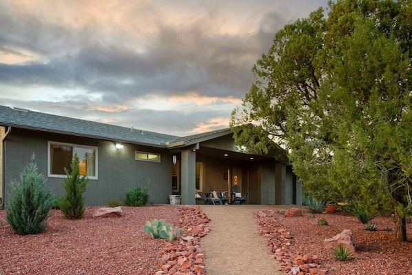 Una casa moderna con un patio cubierto, camino de grava y diversas plantas de desierto.