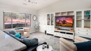 Amplia sala de estar con un televisor de pantalla plana grande, ventilador de techo y vistas a un paisaje rocoso a través de amplios ventanales.