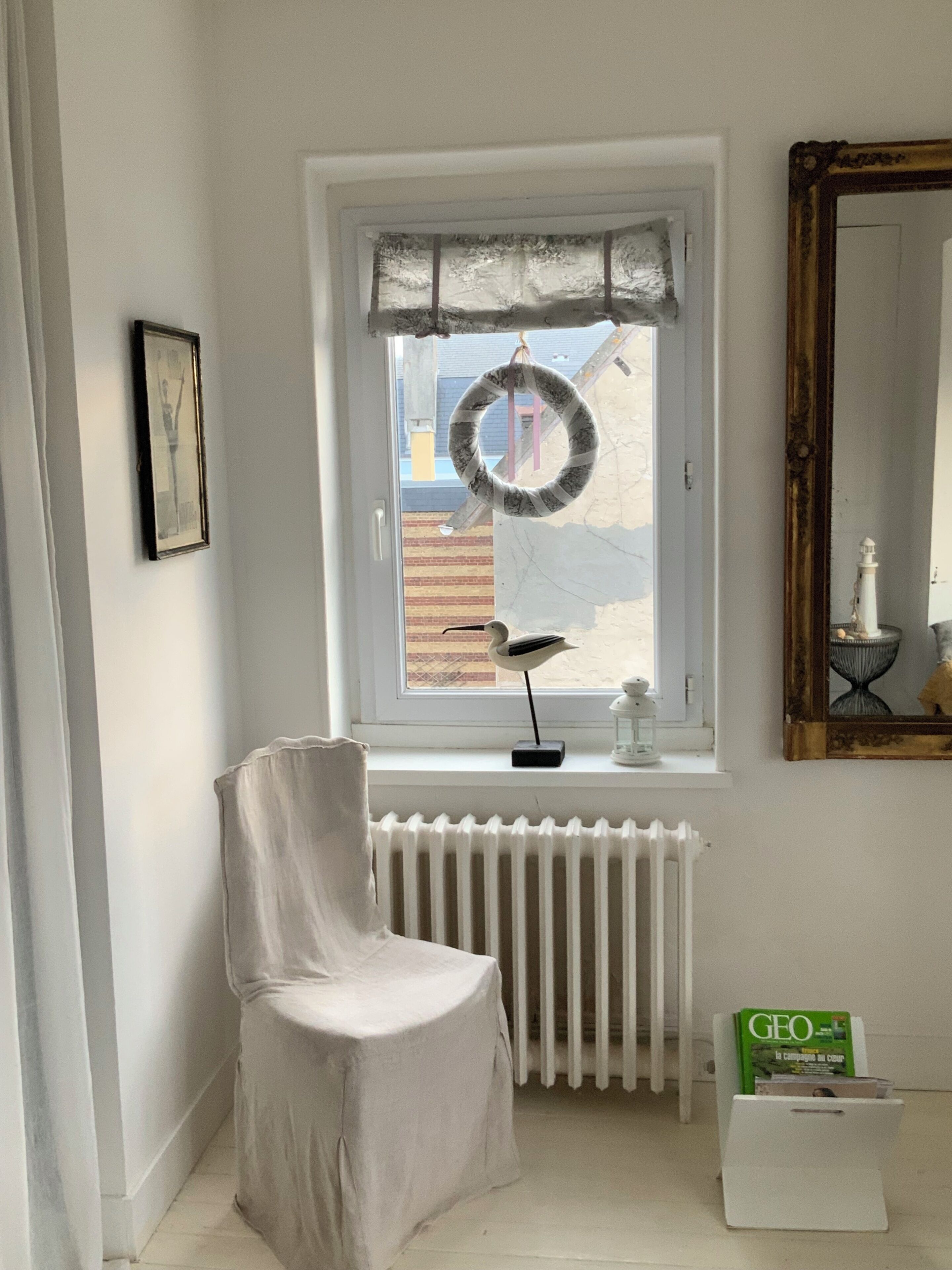 A room with a radiator, a chair, a window with a wreath, a framed picture, and a mirror.