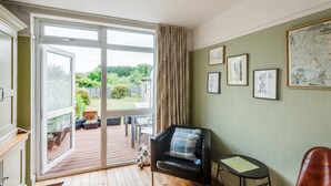 Una habitación con un amplio ventanal corredero que da a una terraza exterior, un sillón de cuero negro con cojín de cuadros, una pequeña mesa redonda y varias obras de arte enmarcadas en la pared.
