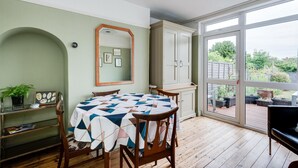 Una habitación con una mesa redonda cubierta por un mantel con estampado geométrico, sillas de madera, un espejo, un armario y vistas al exterior a través de grandes puertas de cristal.