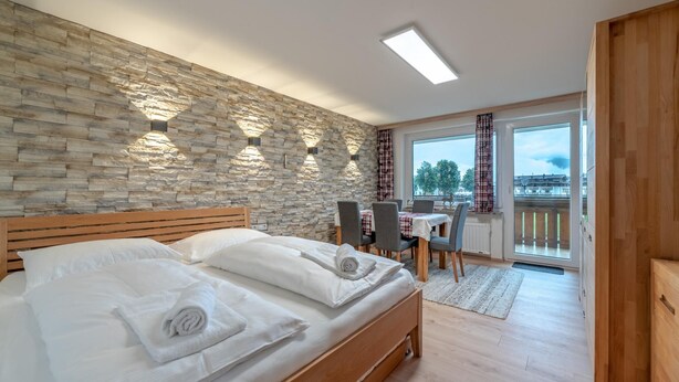 A bedroom with a stone wall, a wooden bed, a dining area with a table and chairs, and a view through a glass door.