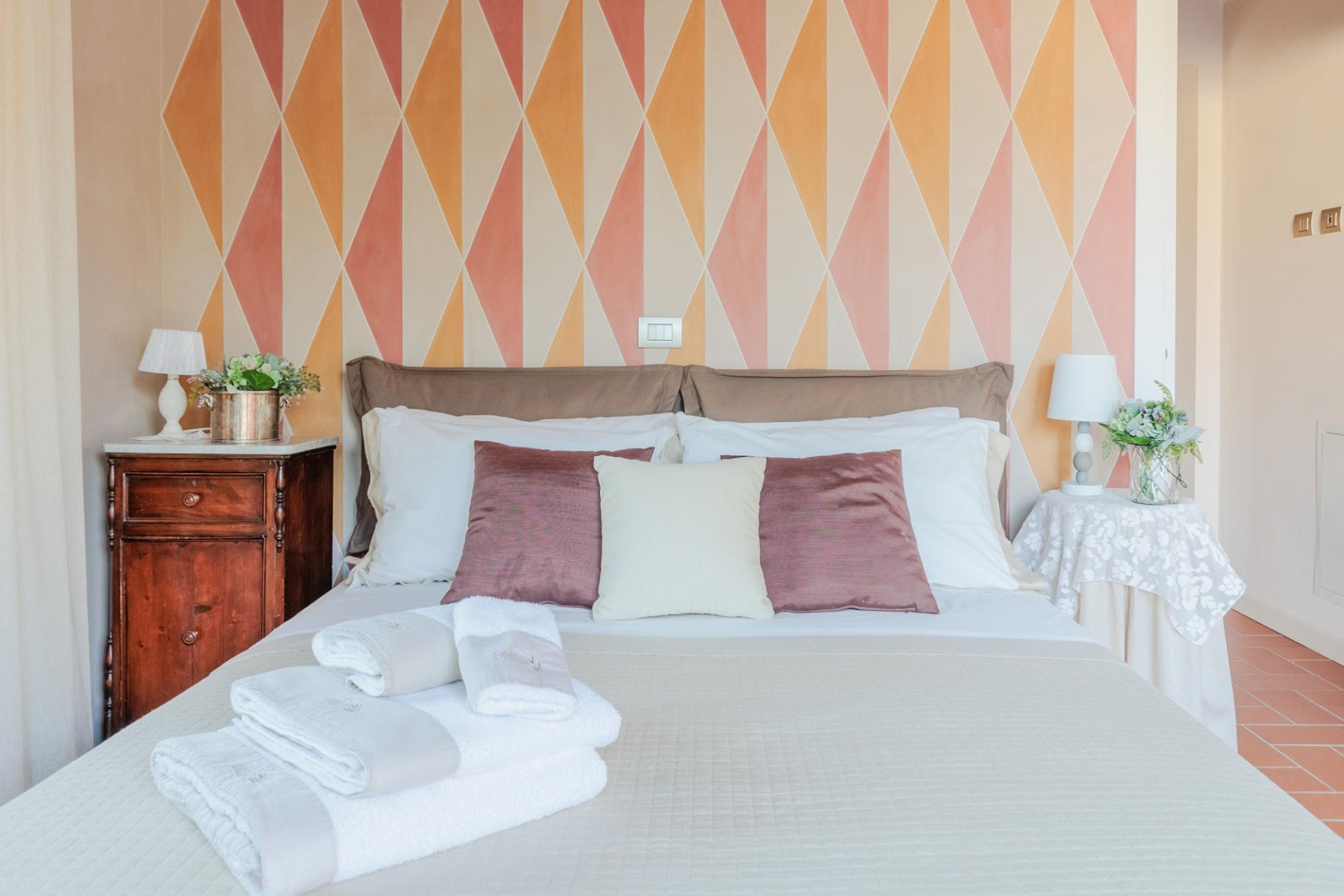 A neatly made bed with white and brown linens, a wooden nightstand with a lamp, and a wall with a geometric pattern.