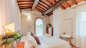 A bedroom with a large bed, a wooden ceiling, a window with curtains, and a small table with towels. - Nonno Giulivo Farmhouse Tuscan Sanctuary (Capannori)