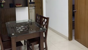 A dining area with a glass-top table and wooden chairs, a wooden cabinet with shelves, a small vase with flowers, and a wooden door. - Captivating 2-bed Apartment in Dhaka (Dhaka)