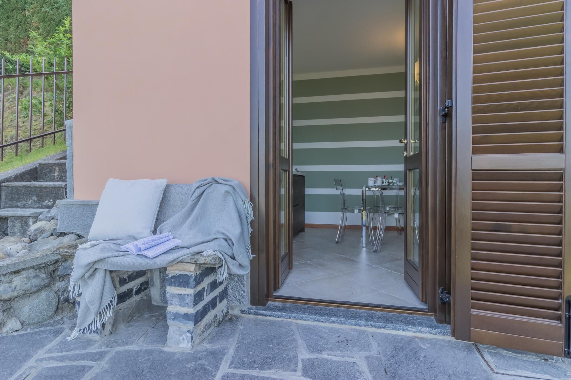 A stone bench with a blanket and pillows, a dining area with a table and chairs, and a sliding glass door leading to a staircase.