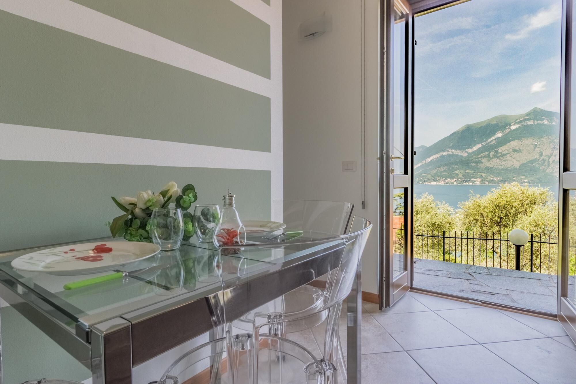 Un espace repas avec une table en verre, des chaises transparentes et une vue sur les montagnes et un lac, visible à travers une porte ouverte.