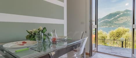 Un espace repas avec une table en verre, des chaises transparentes et une vue sur les montagnes et un lac, visible à travers une porte ouverte.