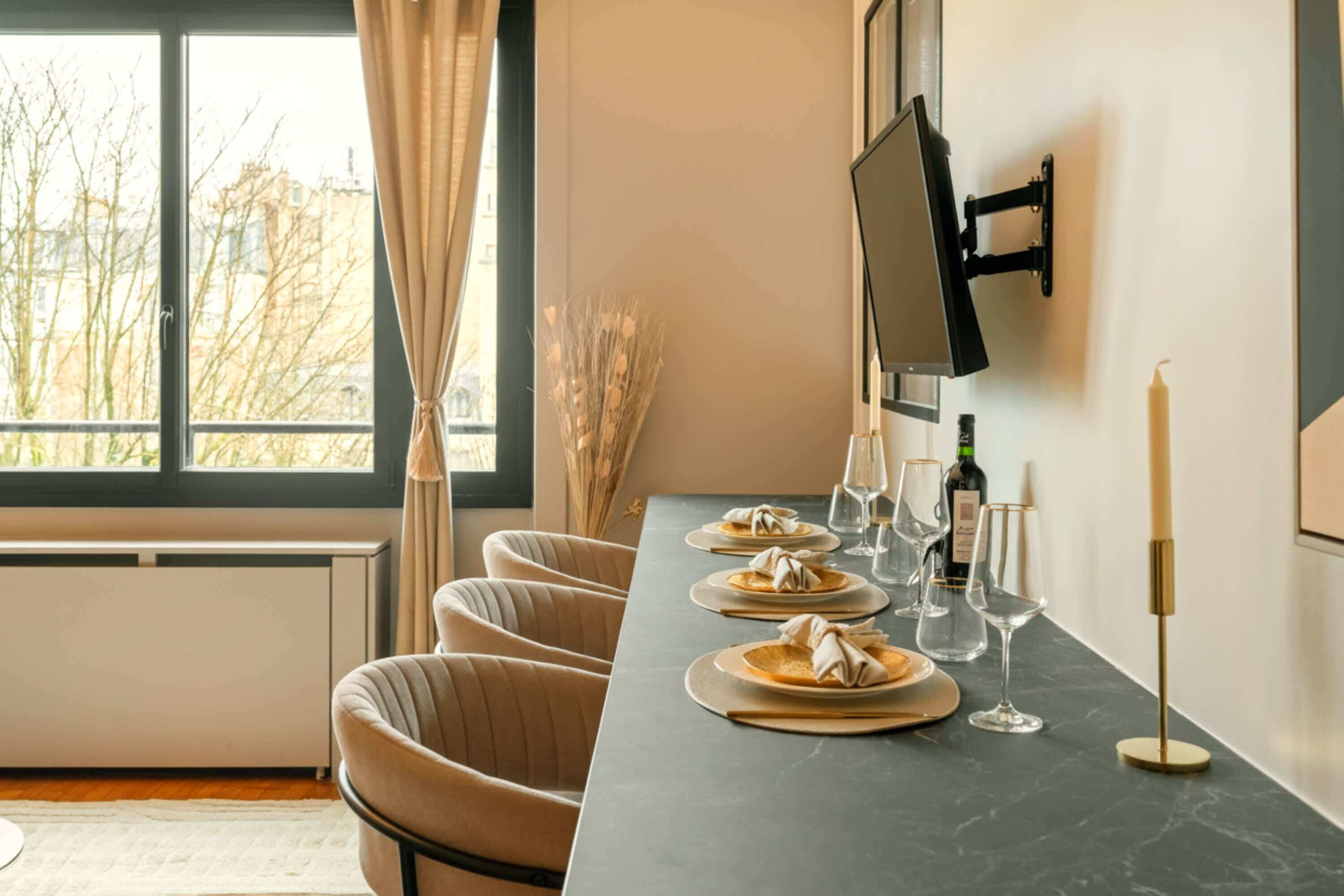 A dining area with a long table, chairs, a flat-screen TV mounted on the wall, and a window with a view of buildings and trees.
