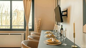 A dining area with a long table, chairs, a flat-screen TV mounted on the wall, and a window with a view of buildings and trees.