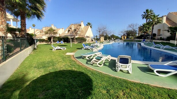 Una piscina con sillones de descanso, un césped bien cuidado y edificios al fondo.