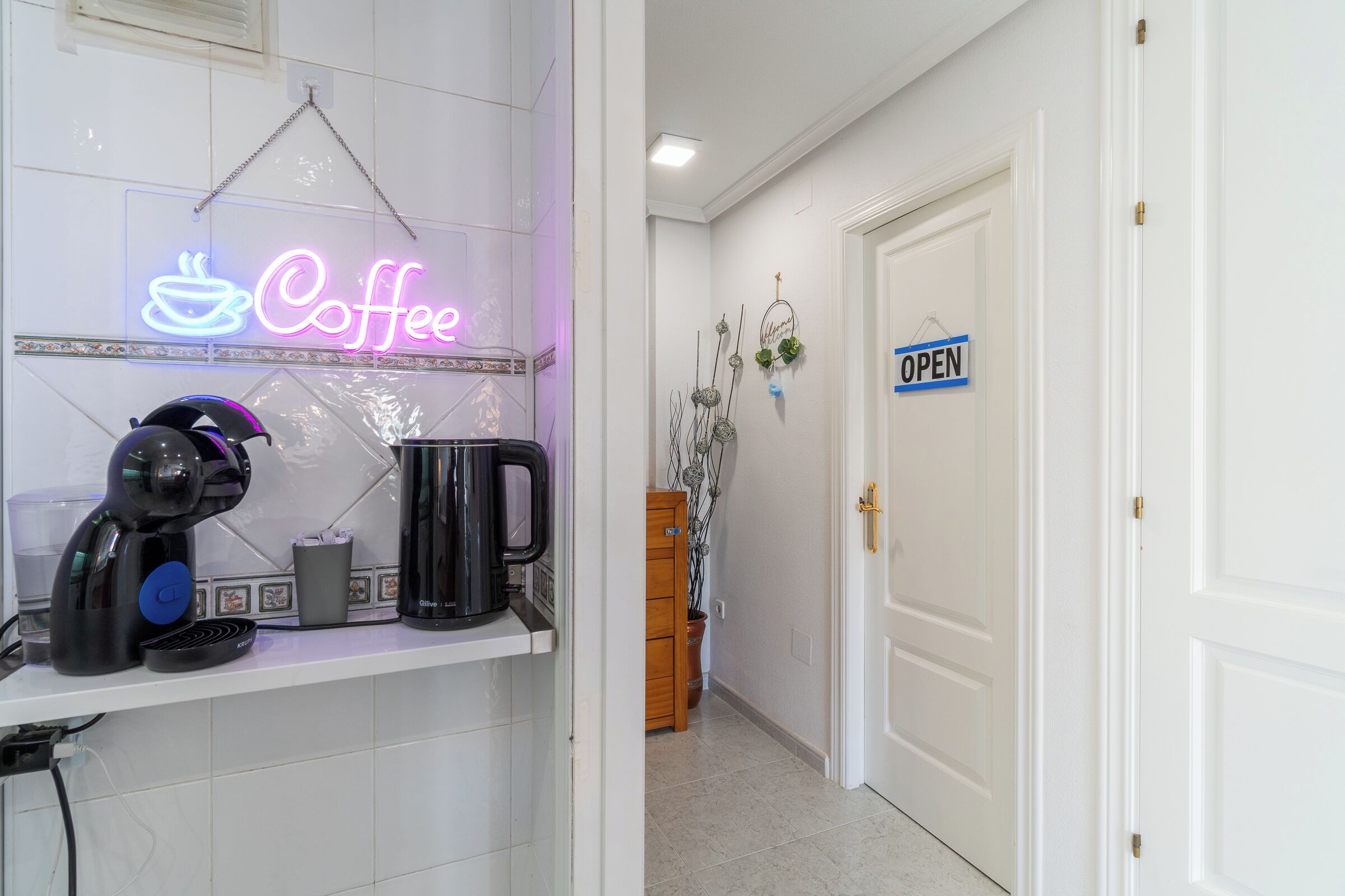 A kitchen area with a neon coffee sign, a coffee maker, and a sign indicating the door is open.