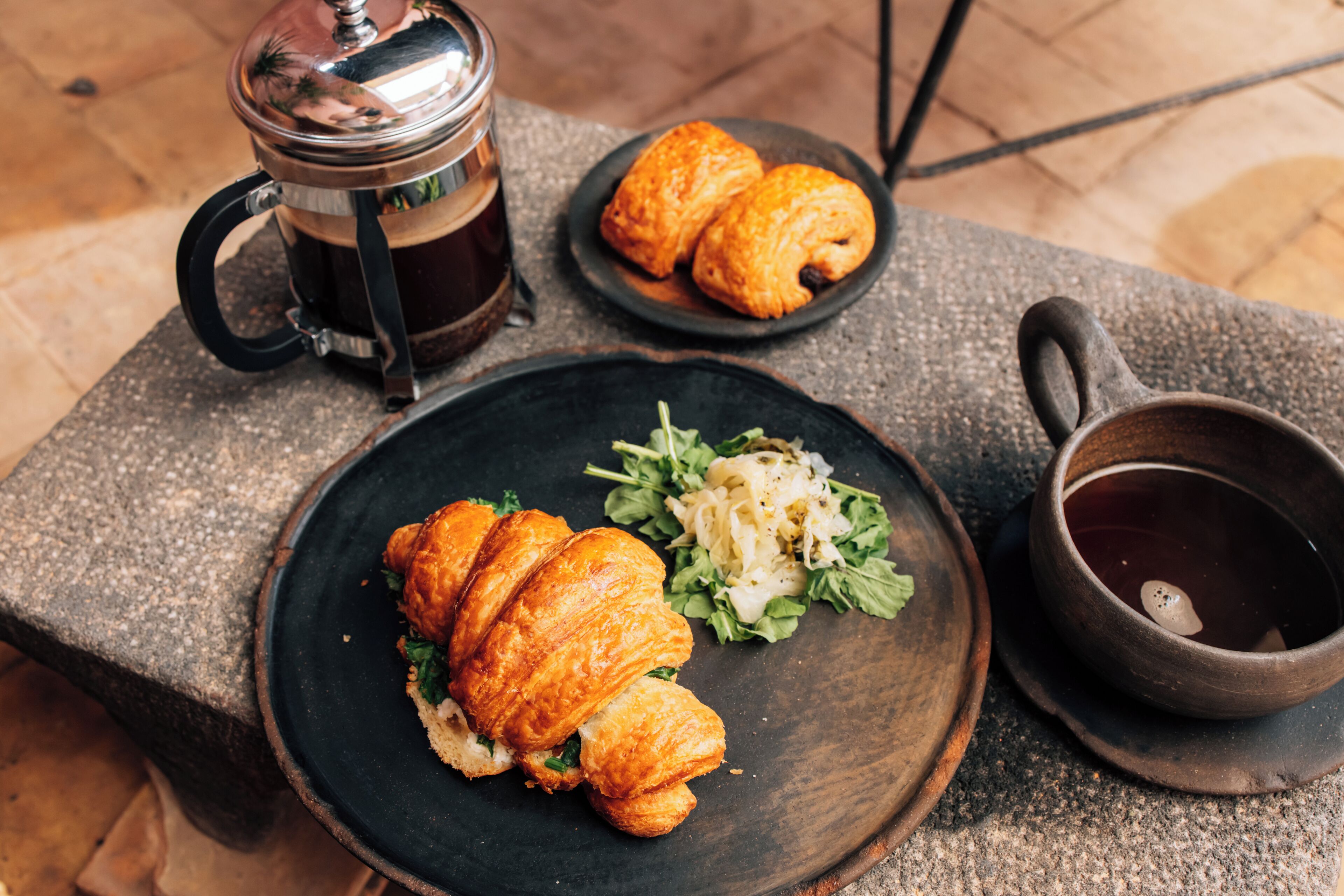 Una mesa de piedra con una fuente de cruasanes, una taza de café y un tazón pequeño de ensalada.