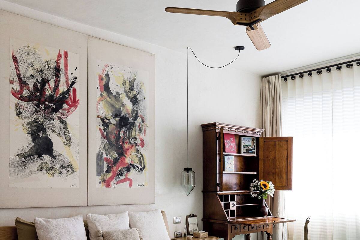A bedroom with a bed, a ceiling fan, abstract art on the wall, a wooden cabinet, and a window with curtains.