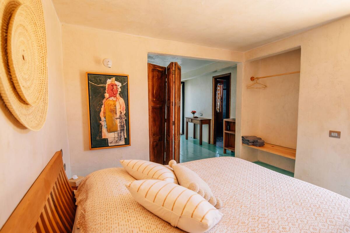 A bedroom with a bed, pillows, a painting, a hat, and a wooden door leading to another room.