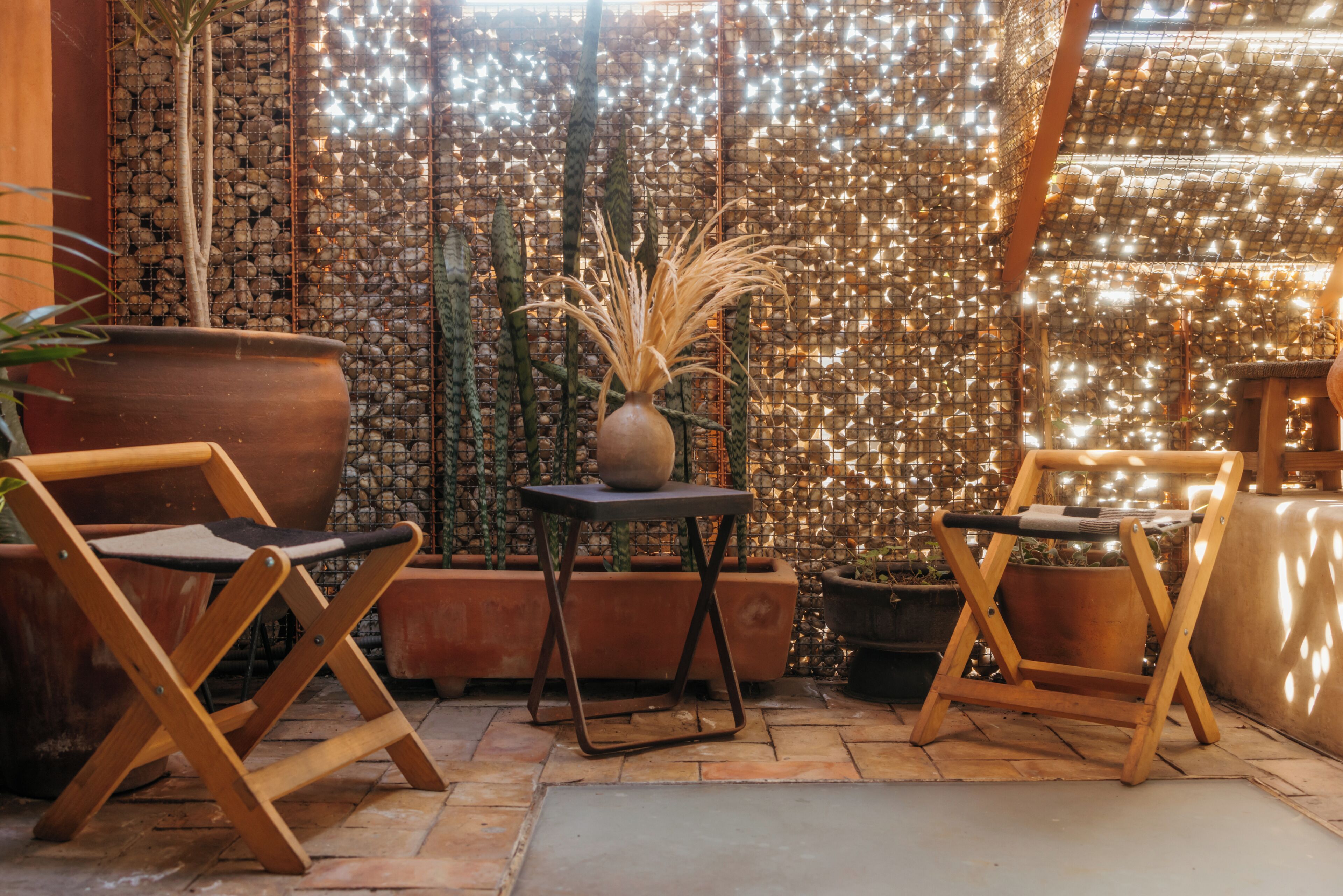 A cozy outdoor seating area with wooden chairs and a table, surrounded by potted plants and a decorative wall.
