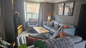Una sala de estar moderna con un sofá gris, una mesa de centro de madera y un sillón gris. Se observa una ventana con cortinas, una lámpara y una planta.
