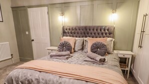 Una cama bien tendida con una colcha estampada y almohadas, dos mesitas de noche con lámparas, y un radiador en la pared.