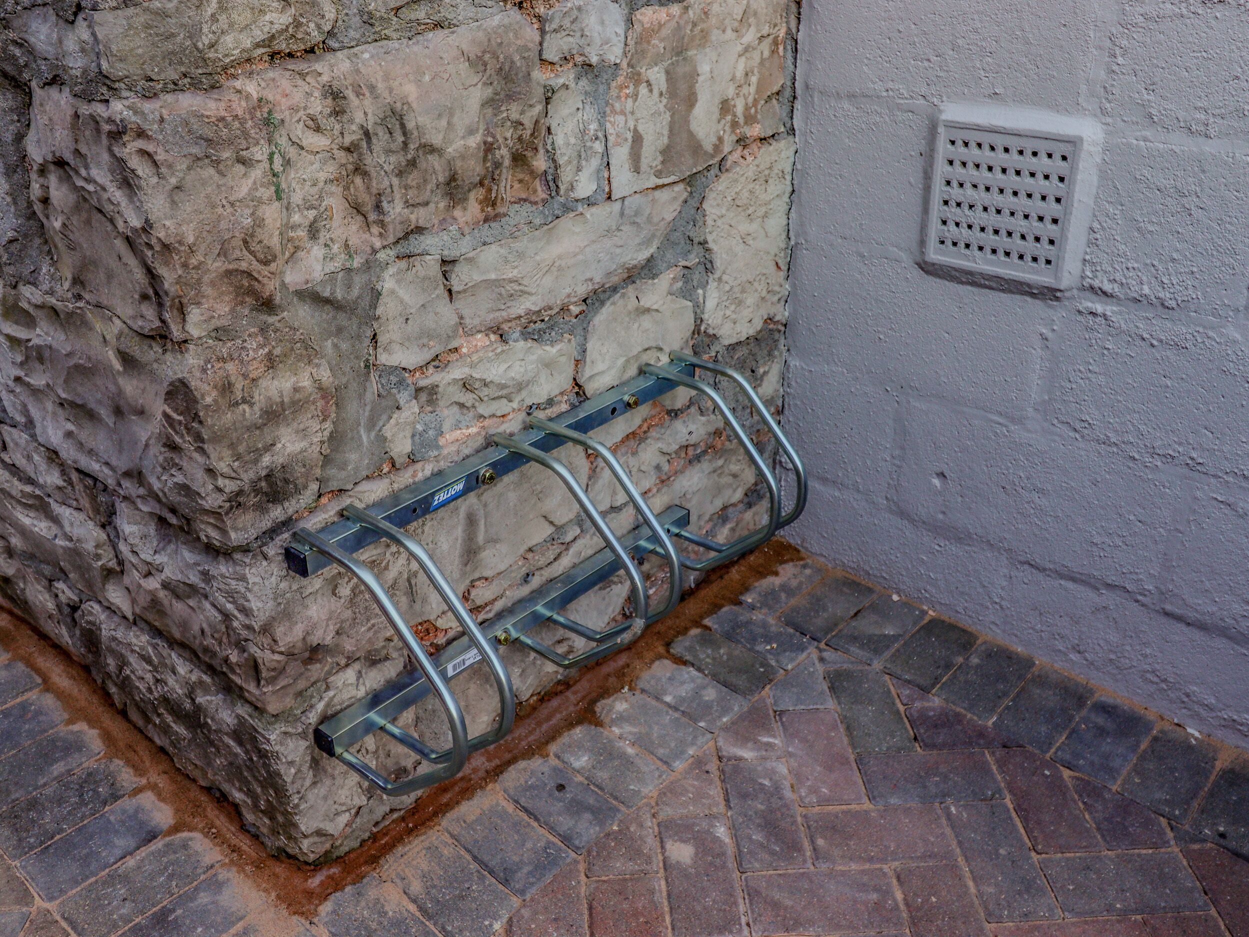 Een metalen rek bevestigd aan een stenen muur, een ventilatieopening in de aangrenzende muur en een tegelvloer.