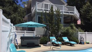 Piscina con sillas de descanso y zona de estar techada, rodeada por una valla blanca y árboles.
