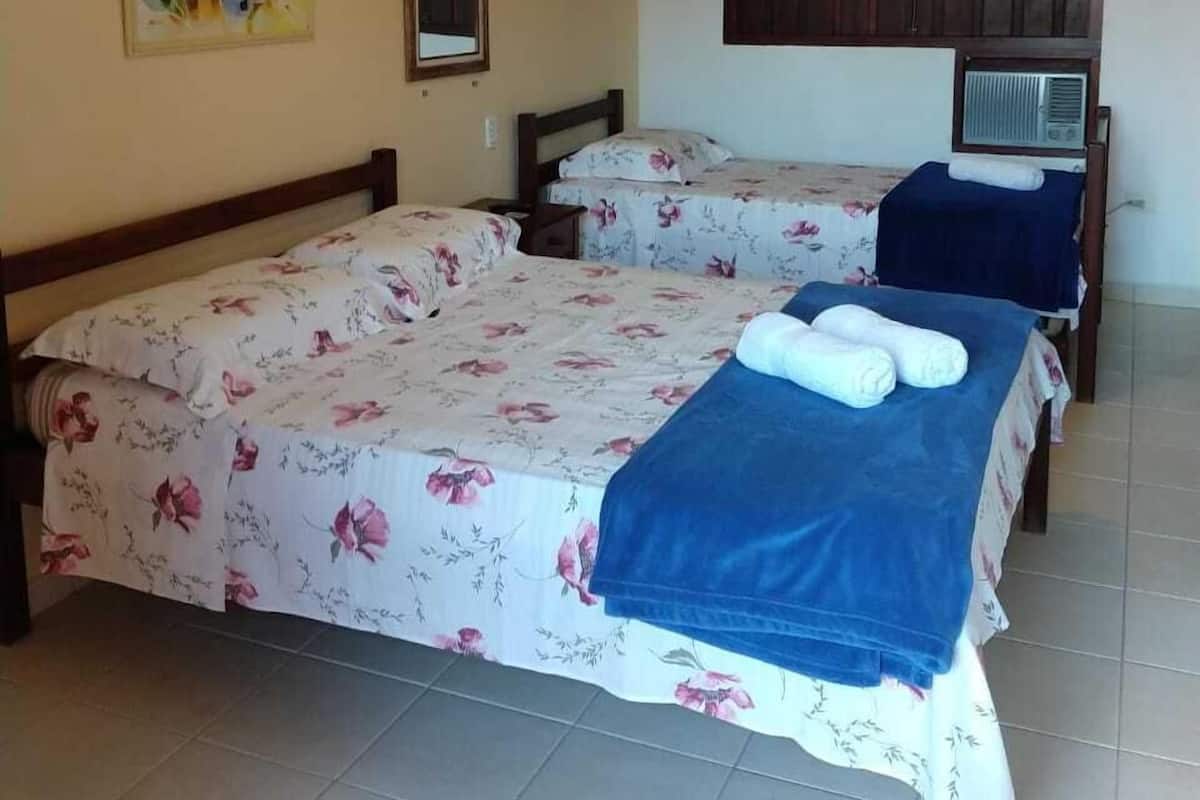 Quarto de hotel com duas camas, um ventilador de teto, uma TV fixada na parede e uma janela com moldura de madeira.