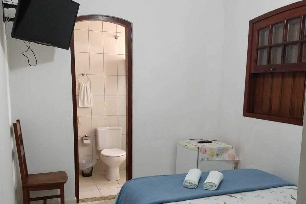 Um quarto com cama, cadeira, televisão, vaso sanitário e janela.