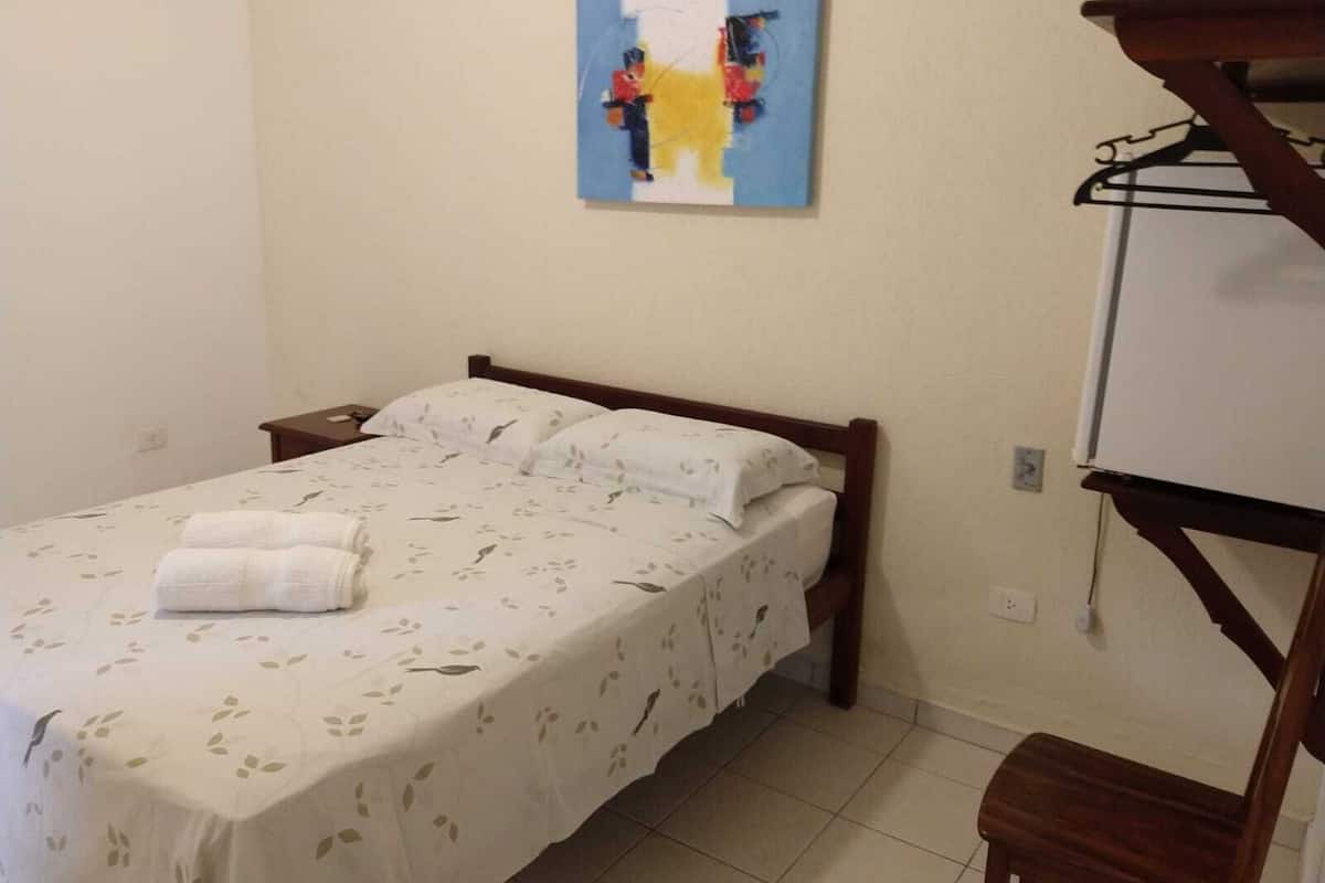Uma cama de solteiro com colcha florida, uma mesa de cabeceira, um quadro pendurado na parede e uma cadeira de madeira.